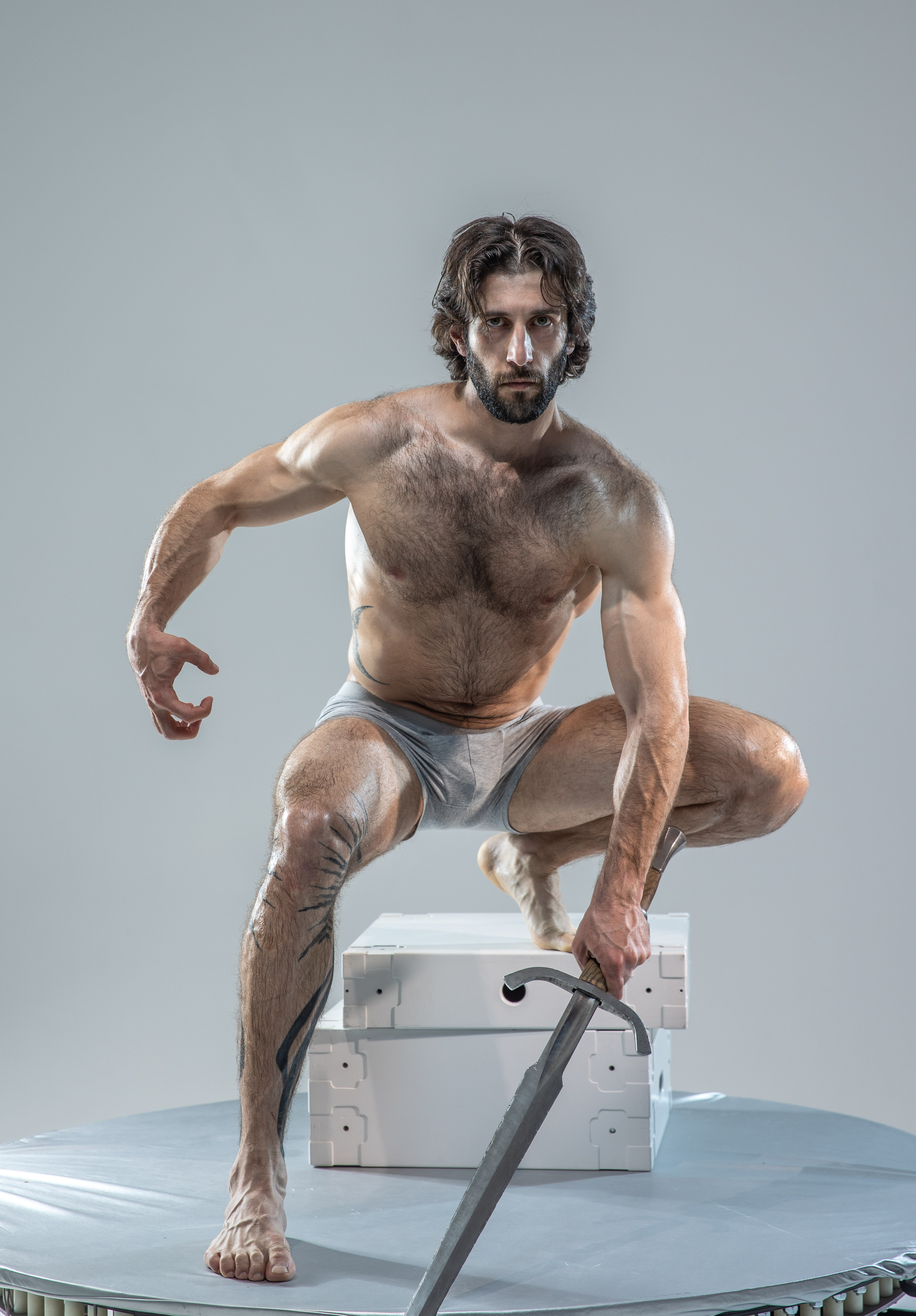 a muscular man with long hair and beard  is posed on a white platform, gripping a sword. he is shirtless, wearing only gray underwear, and has a determined expression on his face.  the background is a solid gray, and the lighting is bright and even.