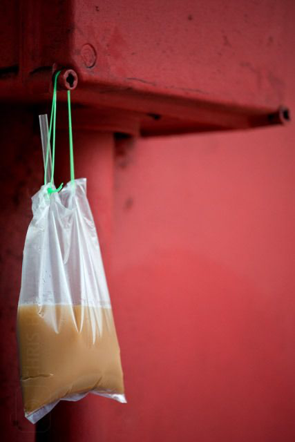 closeup shot of a clear plastic bag filled with a light brown liquid, hanging from a hook on a red wall.  the plastic bag is secured with green string. the background is a solid, slightly textured red wall. the image suggests the bag contains a takeaway drink or food item, possibly street food from southeast asia.