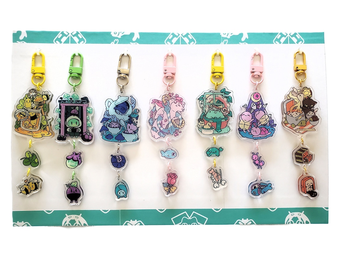 closeup shot showcases a collection of seven uniquely designed acrylic keychains arranged neatly on a teal and white display board. each keychain is a layered charm, featuring an array of whimsical pastelcolored illustrations.  the designs range from a honey bee scene to a tea party setting, an ocean scene, and a picnic. each keychain's design features a top charm and additional smaller charms hanging below on connecting rings. the keychains are displayed against a background that features a repeated logo. the entire image is brightly lit and showcases the detail of the art on each keychain.