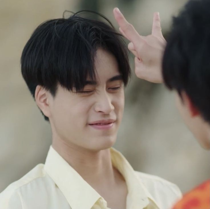 the image shows a closeup portrait of two young men. the focus is on the man in the foreground, who is smiling with his eyes closed as the other man playfully makes a peace sign gesture above his head. the man in focus has short black hair and is wearing a light yellow shirt. the other man is partially visible and wearing a bright orange and yellow shirt. the background is blurry, suggesting a beach or outdoor setting with bright, warm natural light.  the overall tone of the image is happy, playful, and affectionate.