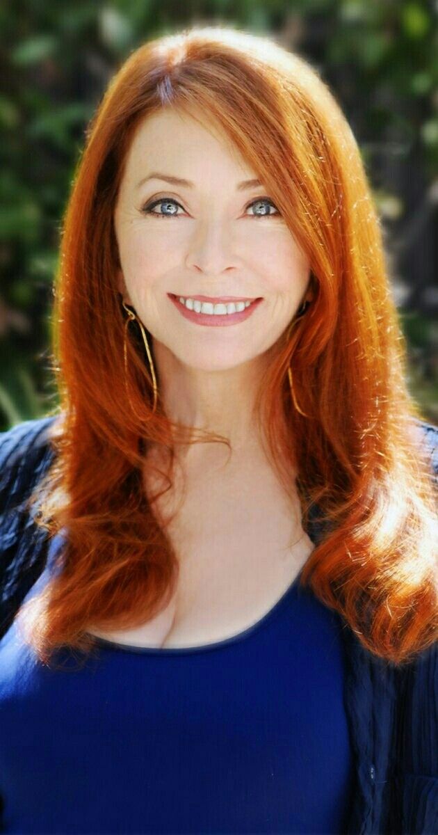 a closeup portrait of a beautiful redhead woman with long flowing hair wearing a blue shirt and smiling at the camera. she is wearing earrings and has a casual and relaxed look. the background is blurry and out of focus, creating a soft and warm feel to the image.