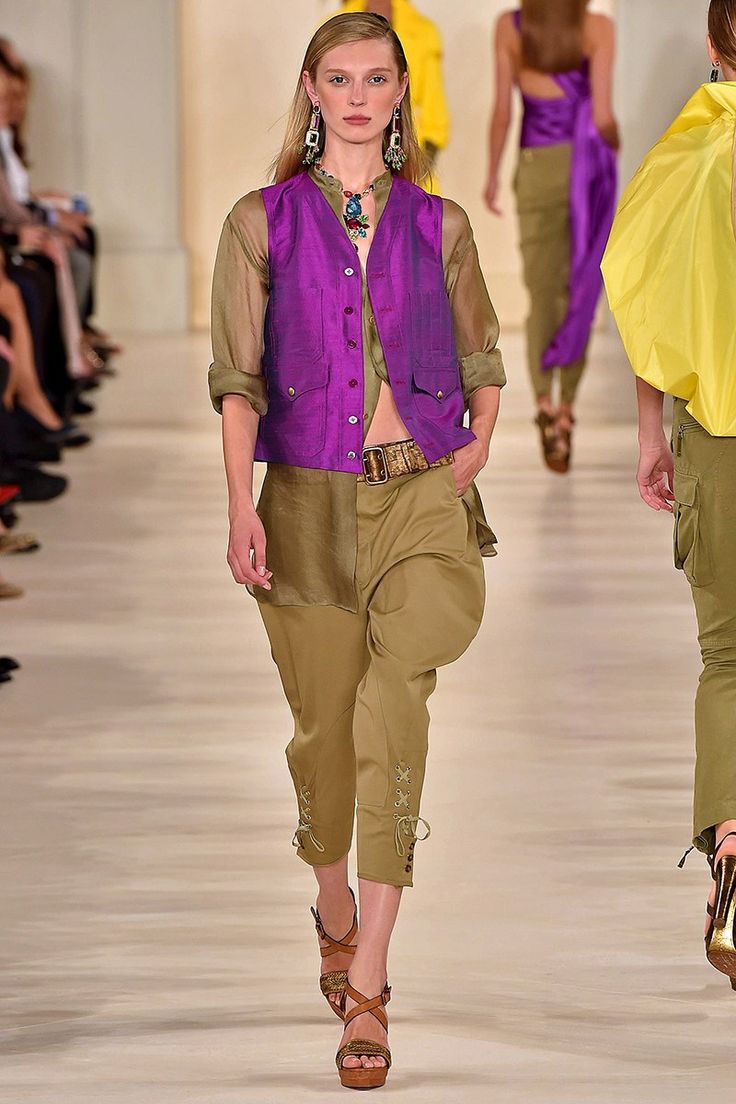 a fashion model walks down a runway wearing a purple silk vest over a khaki shirt and matching pants. the pants are cropped and have laceup details at the bottom. she is wearing brown sandals, a necklace, and earrings. the background shows other models and audience members. the overall style is elegant and sophisticated.