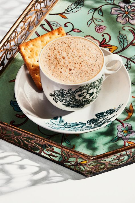 a closeup shot of a cup of teh tarik, a malaysian tea, served with crackers on a floral patterned tray.  the cup and saucer are decorated with a vintagestyle design. the scene is brightly lit, with shadows cast on the surface.  the overall aesthetic is warm and inviting, suggestive of a relaxing moment.