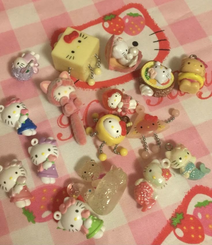 a collection of hello kitty charms and figurines on a pink checkered surface with a hello kitty face and strawberries. the items include various hello kitty themed charms in different designs and poses, some of which appear to be part of jewelry or accessories. the overall aesthetic is kawaii and cute.
