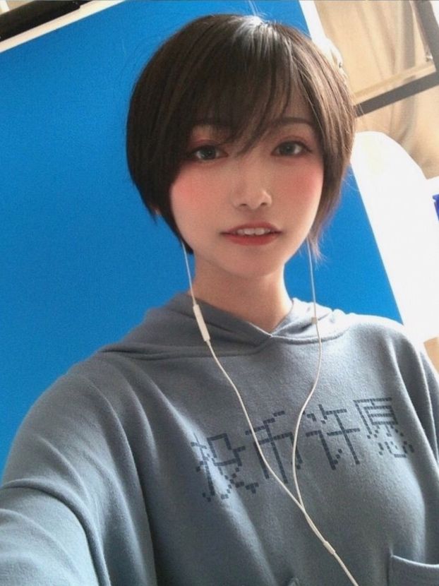 a young asian woman with short hair wearing a blue hoodie is looking at the camera and smiling. she is listening to music with earphones. the background is blue.