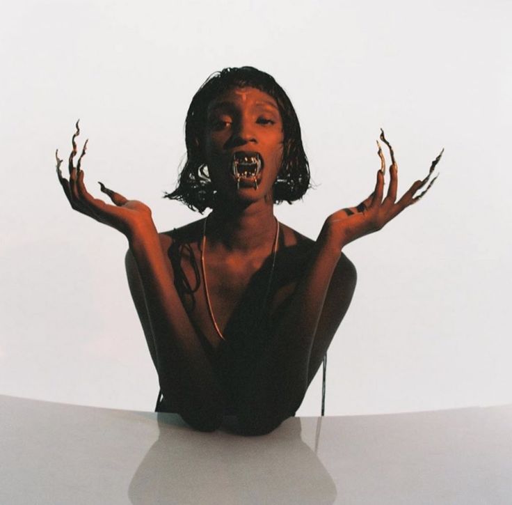 a black woman wearing a black outfit, long sharp nails, and a gold necklace poses in a studio. she has fangs and is staring at the camera with a fierce expression. her  hair is black and short and she is wearing bold makeup.