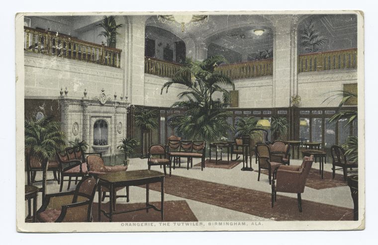 a vintage postcard depicts the interior of the orangerie at the tutwiler hotel in birmingham, alabama. the scene showcases a grand lobby with a balcony, ornate architectural details, lush plants, and various seating arrangements. a decorative fountain is visible, and the overall atmosphere exudes a sense of historical elegance and sophistication.