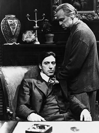 a black and white movie still shows al pacino as michael corleone seated and marlon brando as don corleone standing behind him. pacino is in a pinstripe suit and is seated behind a desk with an ashtray and a lighter. behind them on a shelf are a vase, sculpture, candlestick and pot plant. above the shelf is a framed painting.
