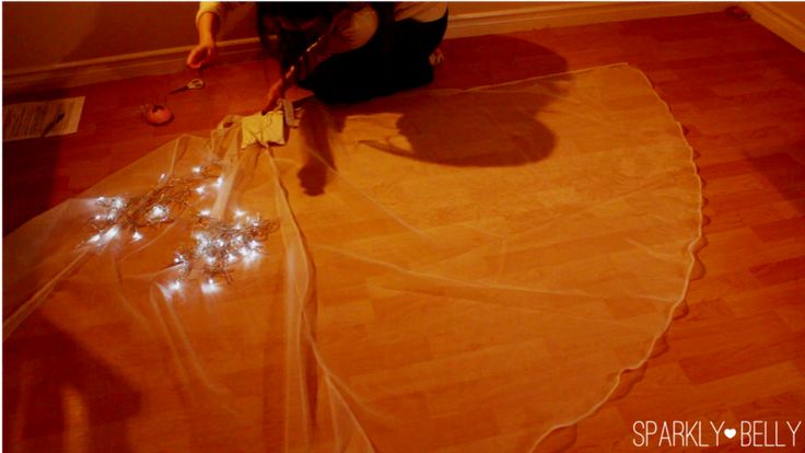 a person is kneeling on a wooden floor, working on a veil that is spread out on the floor. the veil is decorated with lights. there are scissors, thread, and a sewing kit nearby. the image has the text "sparkly <3 belly" in the bottom right corner.