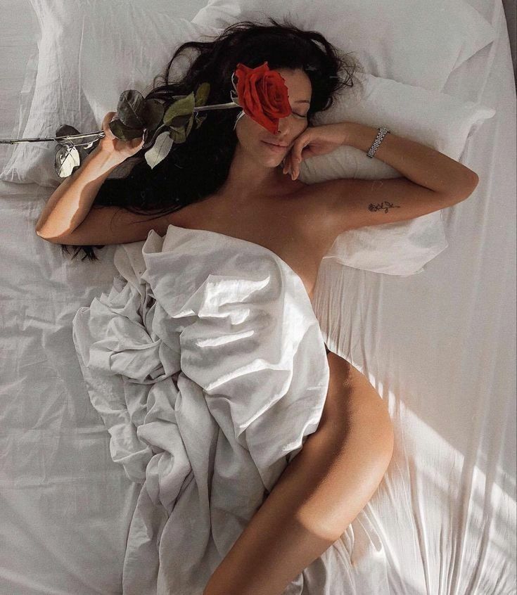 the image shows a young woman with long dark hair partially asleep in a bed.  she is partially covered by white sheets, and a single, longstemmed red rose rests across her face, partially obscuring one eye.  she is wearing a bracelet on her wrist and has a small tattoo on her arm. the scene is softly lit by natural light streaming into the bedroom, giving it a peaceful and sensual ambiance. the overall aesthetic is one of romantic intimacy and relaxation.