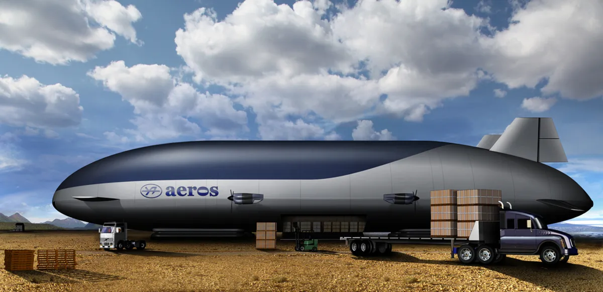 a large, sleek, silver airship with black accents is parked on the ground in a desert environment. the airship, which is labeled "aeros" in the image, has a long, narrow fuselage and two large fins at the rear. there is a semitruck with trailer in front of the airship, and it is loaded with wood crates. the airship is designed for cargo and commercial transport and the image is a digital illustration. the sky is blue with white fluffy clouds.