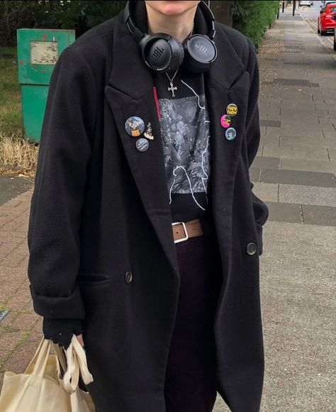 a young woman wearing a black long coat, a black tshirt with badges,  a belt, black headphones and a white tote bag. she's standing on a sidewalk in a city, looking down.