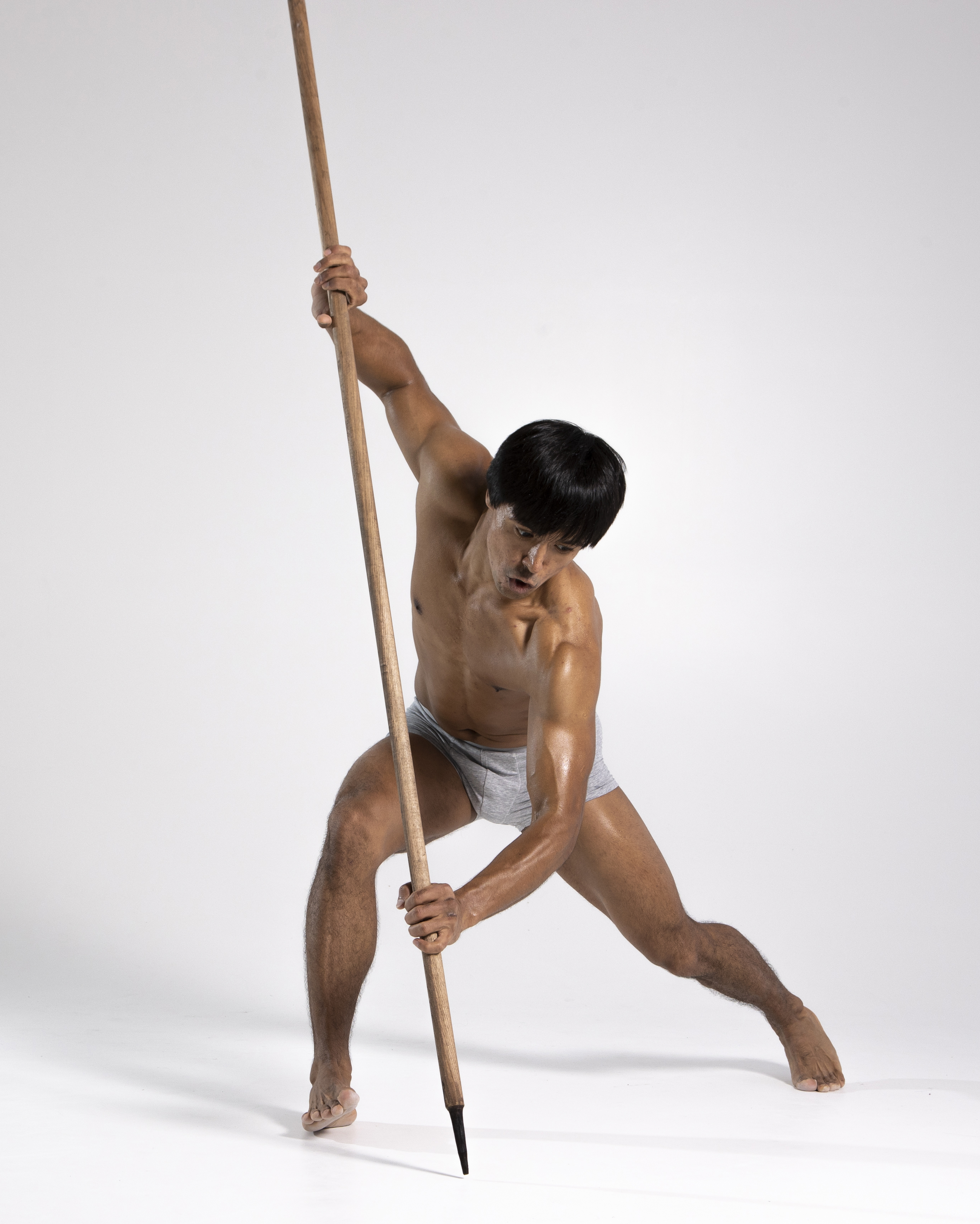 a shirtless, muscular man in gray underwear holds a spear with a black tip in both hands, standing barefoot on a gray background in a studio shot. he is in a wide stance, with one leg forward and bent at the knee, giving the impression of a martial arts warrior or someone posing with a weapon.