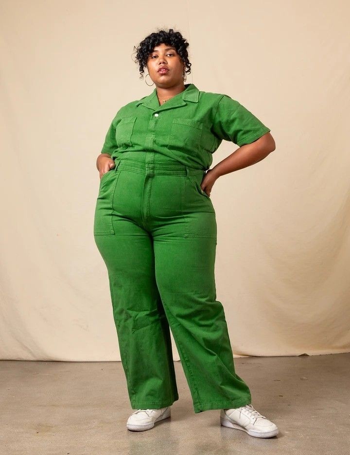 a woman is standing in a studio, wearing a green jumpsuit and white sneakers. she is posing for the camera with her hand on her hip. the jumpsuit is a collared, shortsleeved style with a front zipper and multiple pockets. the background is a cream color, and the lighting is soft and natural.