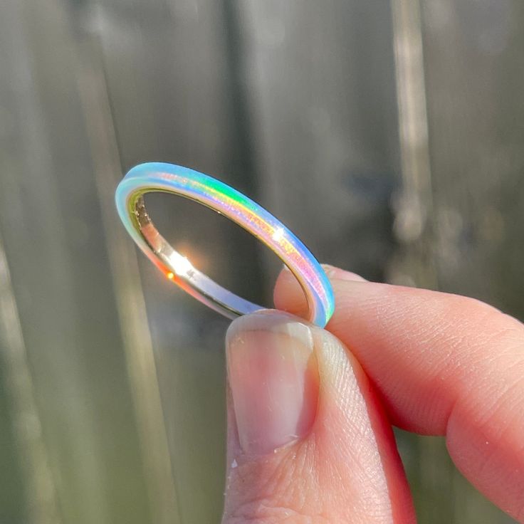 a close up view of an iridescent ring held between two fingers. the ring appears to be made of opal and has a rainbow effect. the ring is set on a gold band.  the fingers are white and the background is blurry.