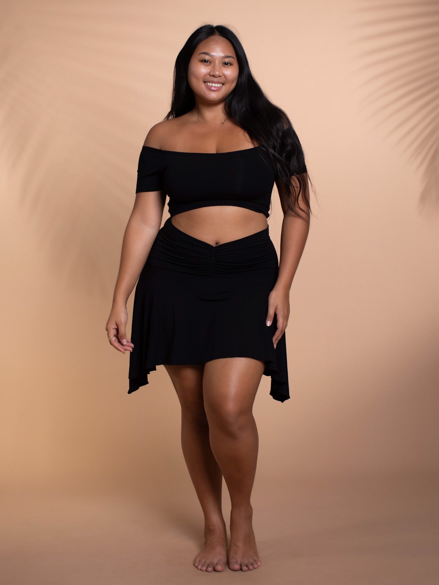 a fulllength studio shot of a smiling plussize woman confidently posing. she is wearing a black offtheshoulder crop top and an asymmetrical black mini skirt. the background is a neutral tan color, and the overall aesthetic is casual and summery.  the image projects confidence and body positivity.