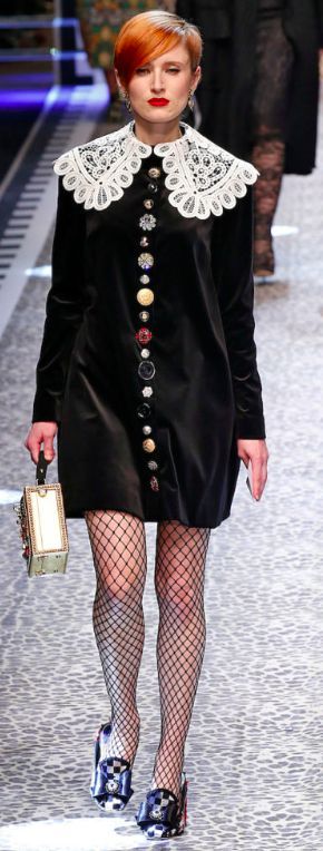 a runway model with short, bright red hair is showcased wearing a black velvet dress adorned with a large white lace collar and a vertical line of colorful buttons down the center.  she's also wearing fishnet stockings and patterned shoes. a small, ornate handbag hangs from her arm. the background suggests a runway setting with a patterned floor. the overall style is elegant and high fashion.
