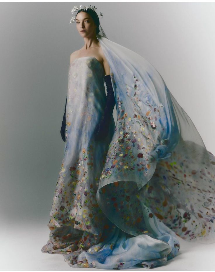 a model in a beautiful, flowing white and blue wedding dress with a floral pattern and long veil. the dress is strapless and has a flowing skirt. the model is posing with her arms down at her sides. the background is a light gray.