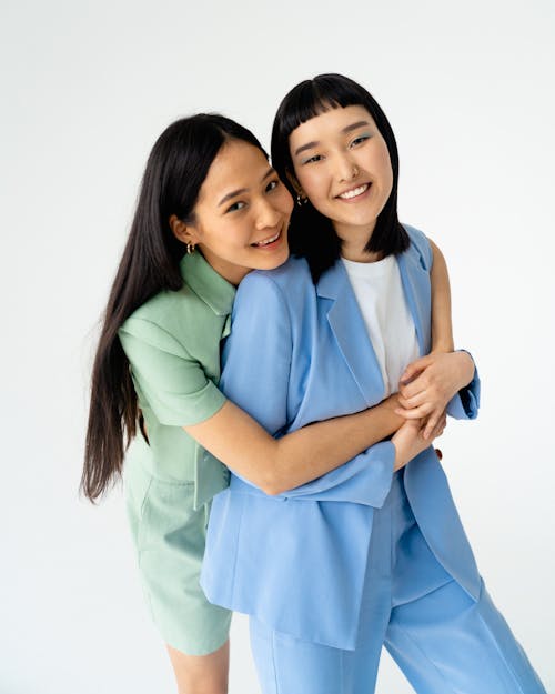 the image shows two young women, one wearing a light blue suit and the other in a light green shorts and shirt set, embracing each other warmly. they are both smiling and appear happy. the setting is a simple white studio background.  the image focuses on their affection and close relationship. the color palette is soft and pastel.
