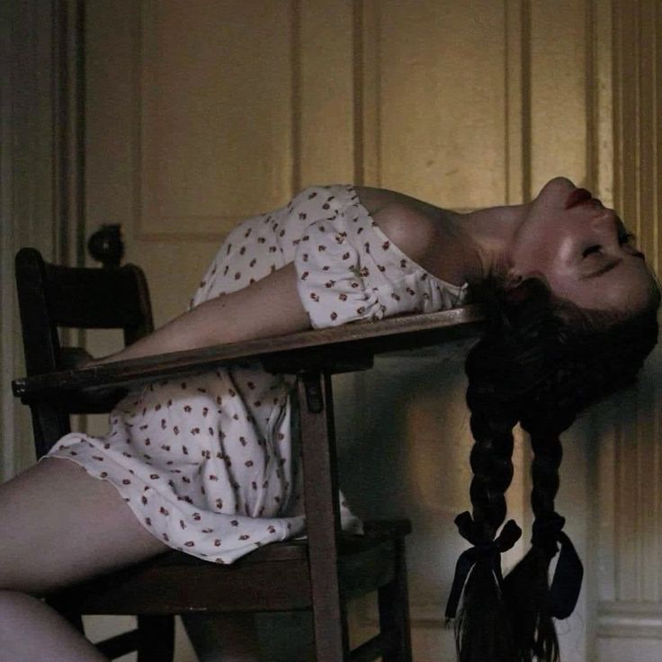 a woman with dark hair in braids with black ribbons is lying backwards over a wooden desk and chair. she is wearing a white dress with a small floral pattern and her head is tilted back with her eyes closed. she has red lipstick on. the background appears to be an old room with wooden panels.