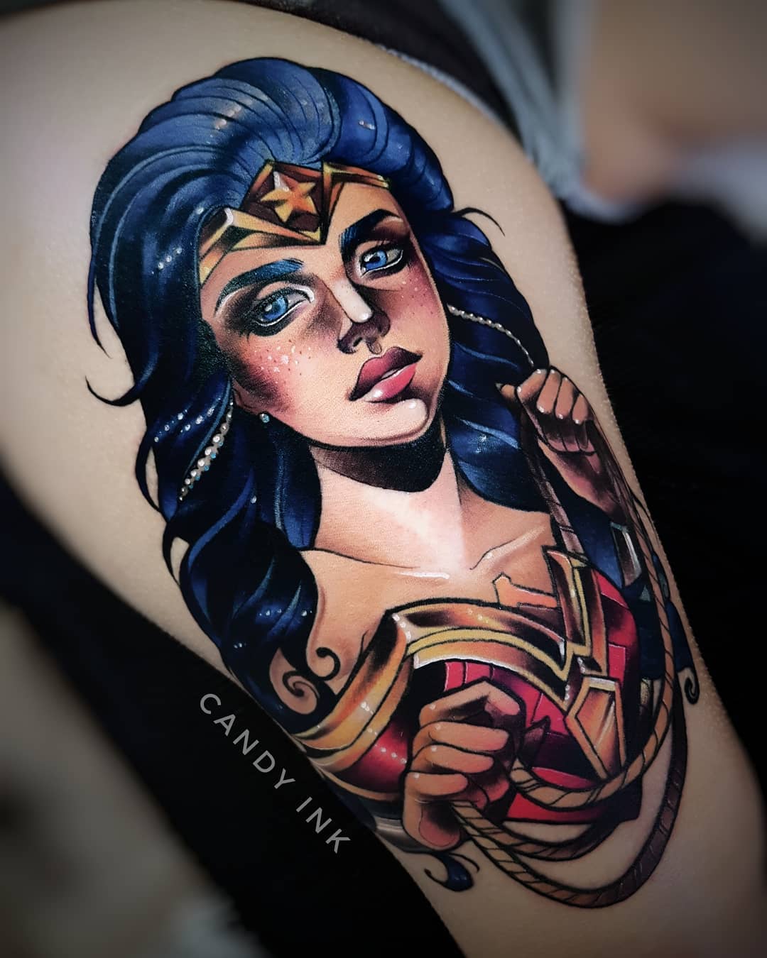 a neo traditional style tattoo of wonder woman on someone's upper thigh. the tattoo has bold black lines and shading, with areas of vibrant color. the background of the tattoo is the person's skin.