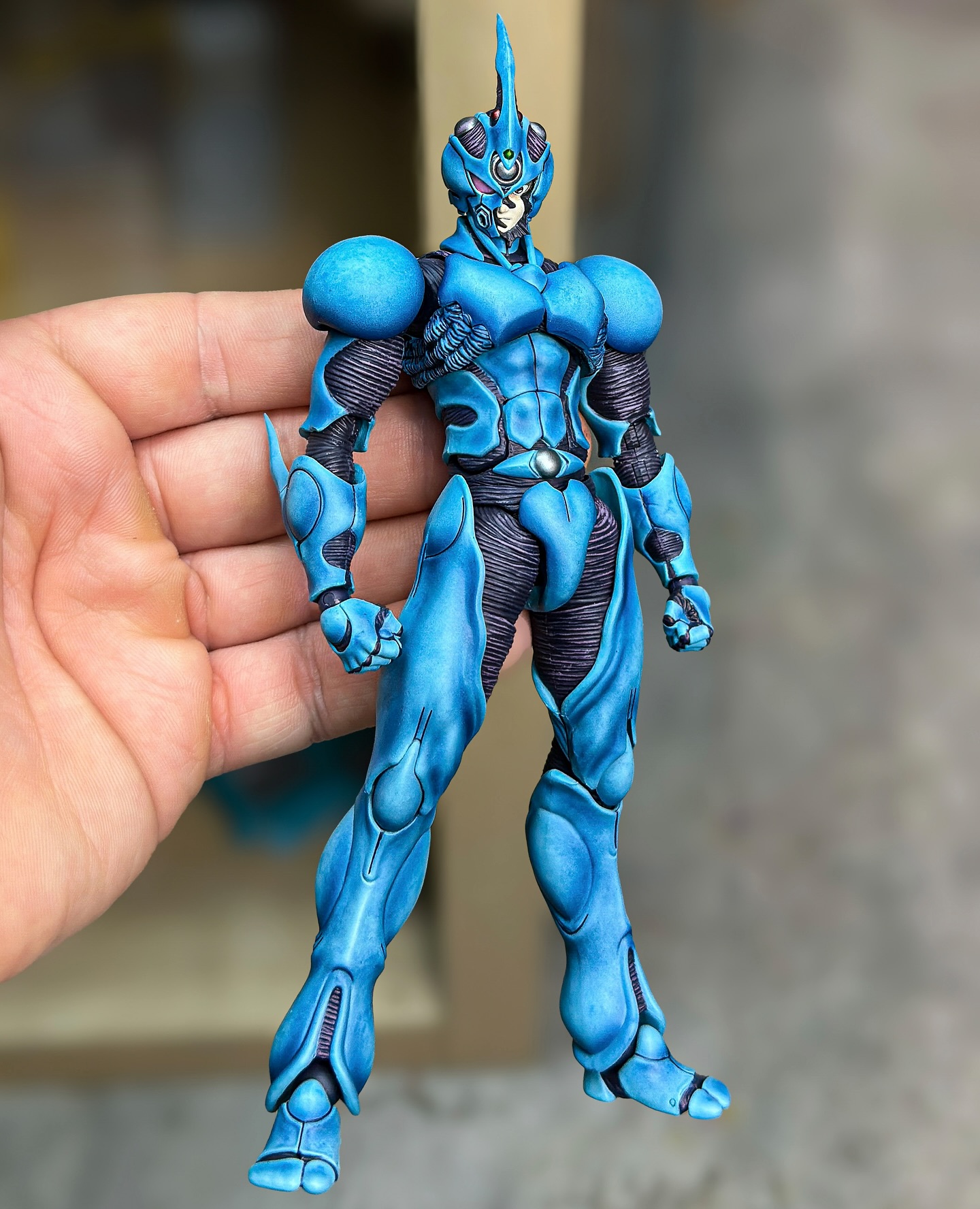 the image shows a blue, armored action figure being held in a person's hand. the figure has a humanoid shape with detailed armor plating, a headpiece with a hornlike projection, and articulated joints. the color scheme is predominantly blue with dark accents highlighting the underlying structure. it is a collectible figure, likely from a manga or anime series.