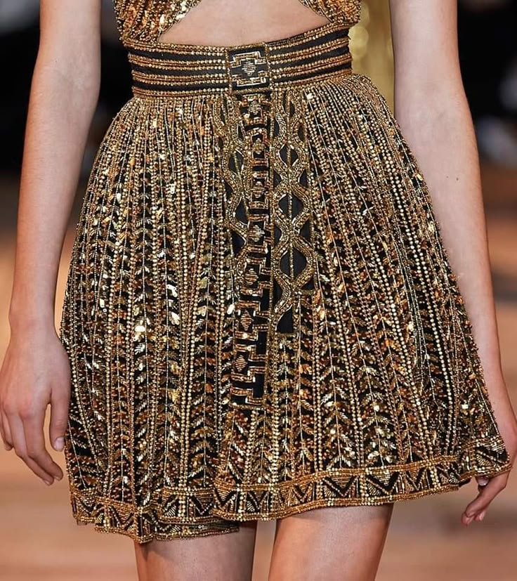 a closeup shot of a woman wearing a short, gold dress heavily embellished with beads and sequins. the dress features a black belt with a decorative buckle and intricate gold patterns on a black base. the dress has a detailed hemline.