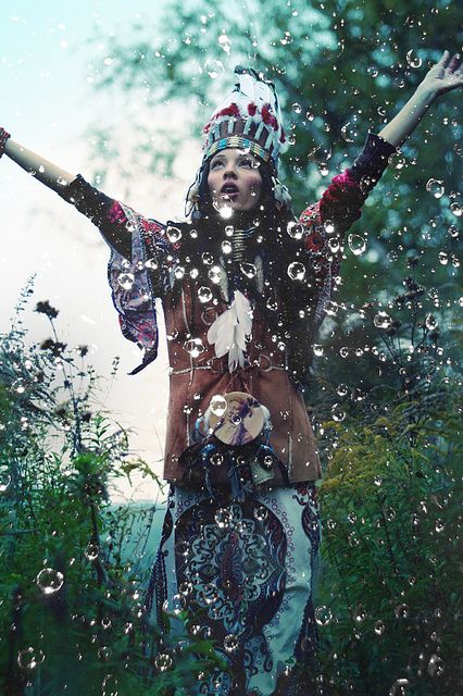 a woman wearing a native americaninspired headdress with feathers and beads, and a long dress with intricate designs, stands in a forest with her arms raised, bathed in a mystical rain of water drops. her expression is serene and ethereal, creating a captivating portrait of a spiritual connection to nature. the backdrop of lush greenery and falling water adds to the enchanting and magical atmosphere of the image.