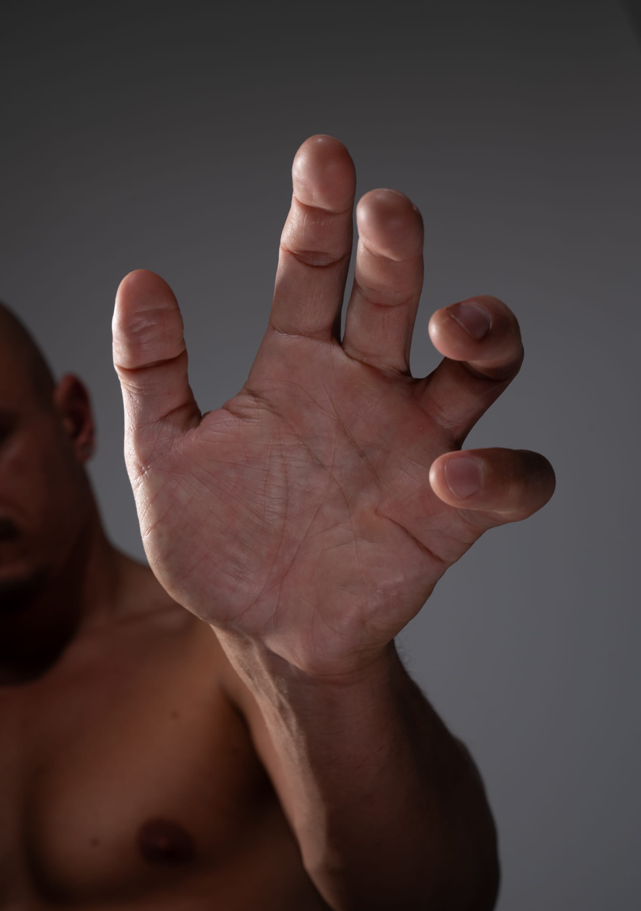 the image shows a closeup of a man's hand and arm. the hand is outstretched with the fingers slightly curled. the man is shirtless and his skin is pale. the background is a dark gray. the lighting is dramatic and creates shadows on the man's hand and body.
