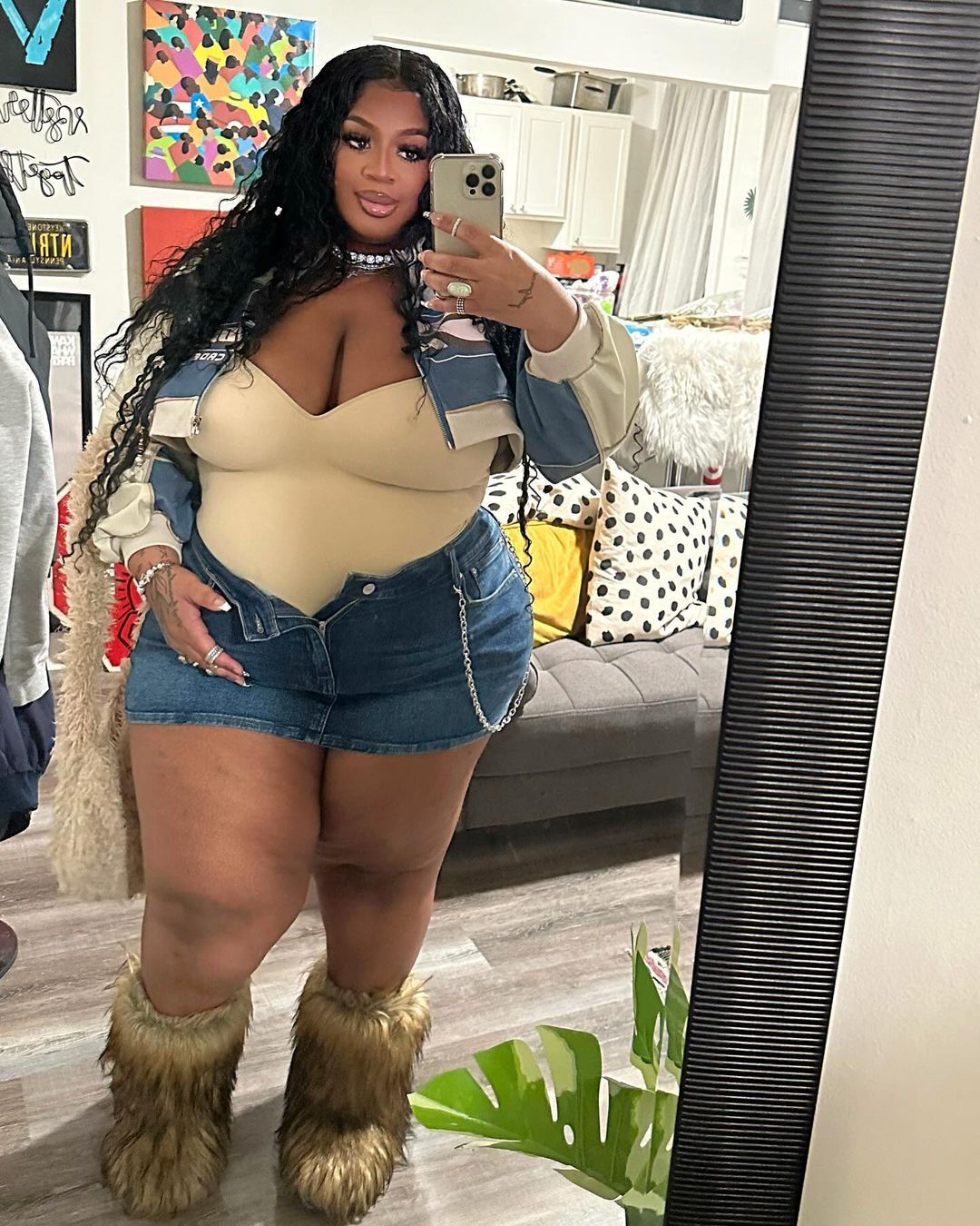 a young black woman in a stylish outfit is taking a selfie in a mirror. she is wearing a crop top, denim skirt, and fur boots, along with a jacket and jewelry.  the image is shot indoors, with a couch, a plant, and a mirror in the background.
