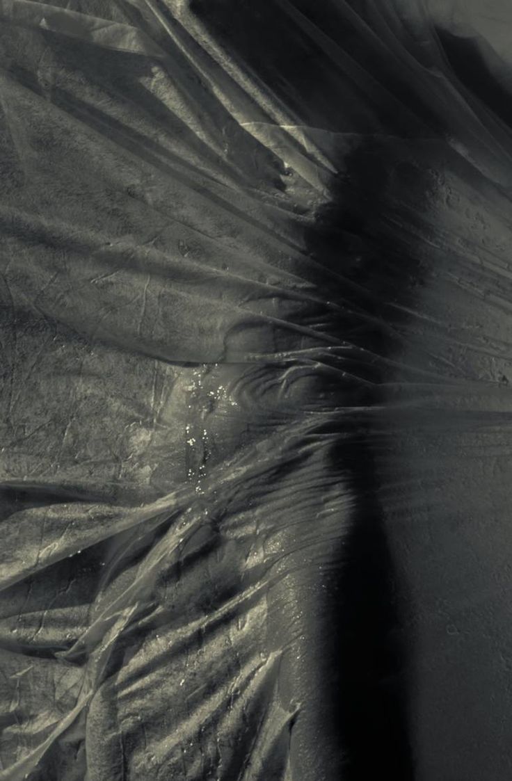 he image is a closeup shot of a plastic sheet draped over a surface. the plastic is textured and shows folds and wrinkles.  water droplets are visible on the plastic.  strong shadows are cast across the scene, creating a dark and moody atmosphere. the overall feel is abstract and artistic, with an emphasis on texture and light. the image is in monochrome (grayscale).