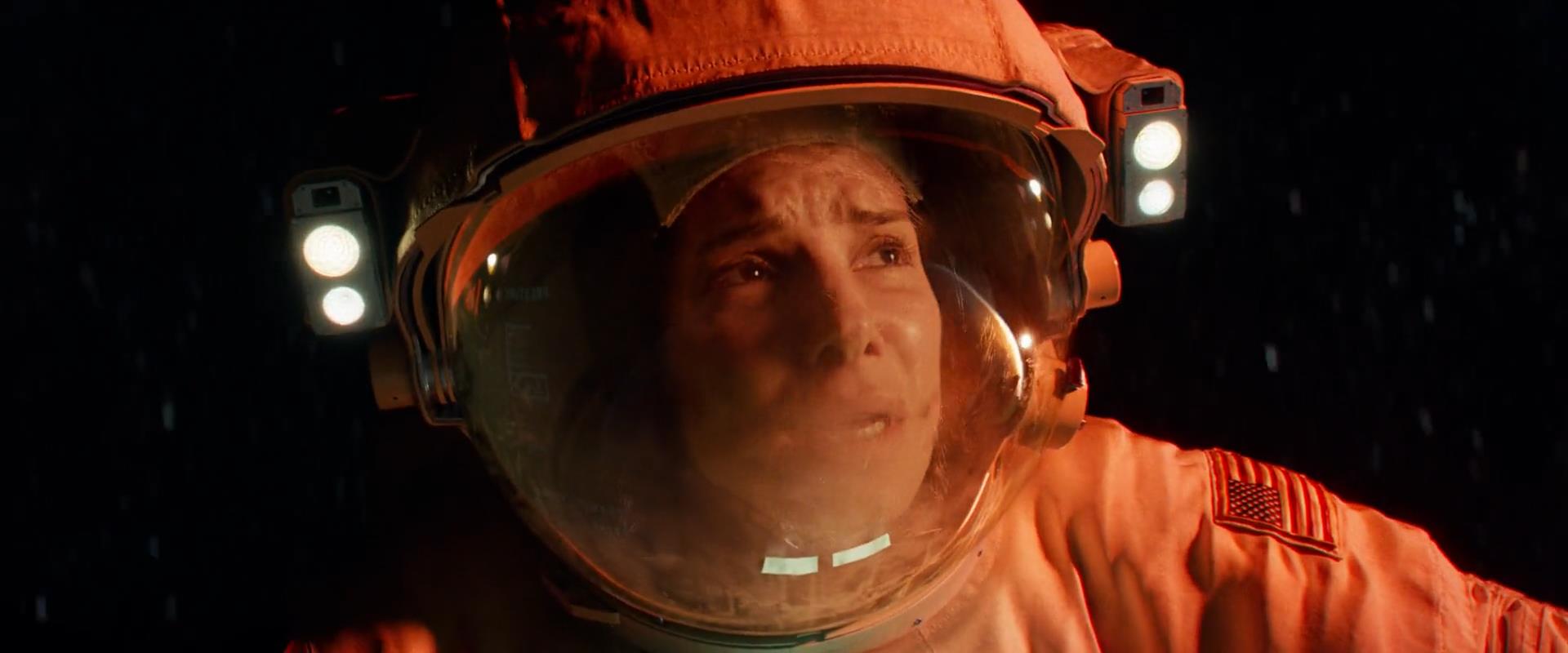 the image shows a closeup of a woman astronaut's face behind the visor of her helmet. the woman appears to be in distress, with a fearful or anxious expression. the lighting is dramatic, with a redorange hue suggesting an emergency situation. the background is the blackness of space, emphasizing the character's isolation.