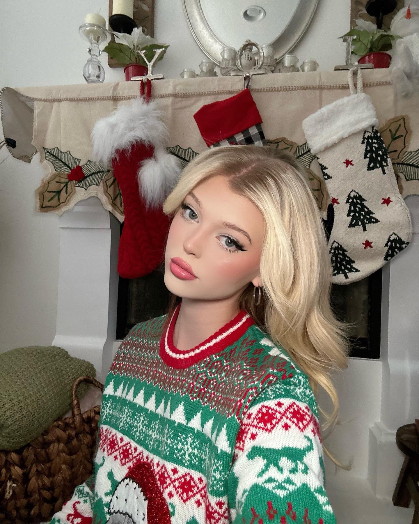 a young woman with blonde hair is wearing a christmas sweater in front of a fireplace with stockings. the room is decorated for the holidays. the woman has makeup on and is looking at the camera. the sweater has a fair isle pattern and is green and red.
