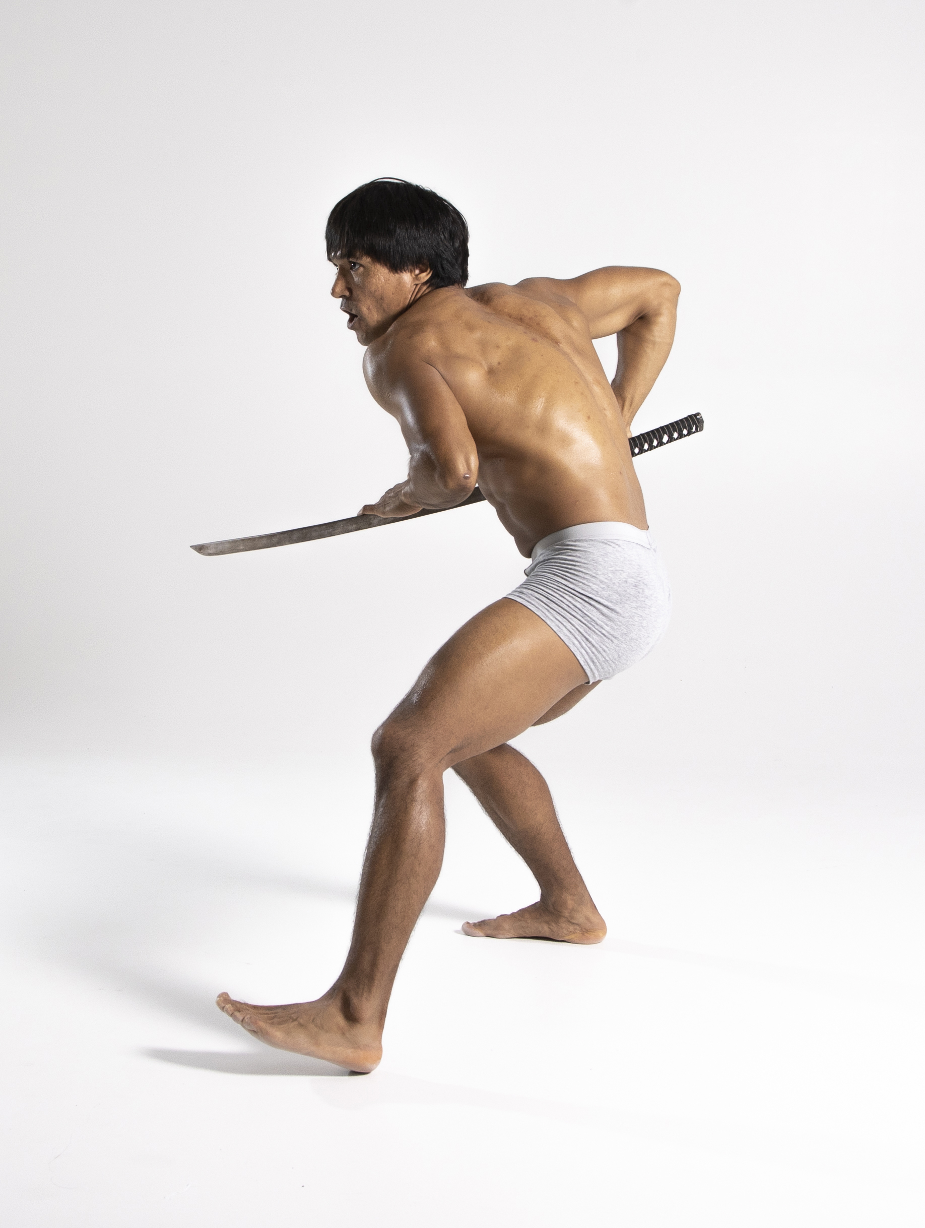 a shirtless, muscular man with a katana is posed defensively in a studio shot against a white background. he is wearing gray underwear and is barefoot. his tense expression and defensive stance suggest a martial arts or combat scenario.
