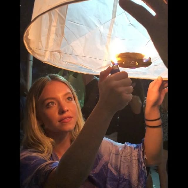 the image shows a young blonde woman,  closeup from the chest up,  in the process of lighting a large white sky lantern.  she holds a lighter to the fuel source inside the lantern while her other hand supports the lantern's structure.  another hand, likely belonging to someone else, is visible in the upper right corner, assisting or observing. the background is dark and blurry, suggesting a nighttime setting, possibly at a festival or cultural event where sky lanterns are being released. the woman is wearing a lightcolored, patterned top. the focus is primarily on the woman and the lantern. the overall atmosphere is peaceful and somewhat magical due to the soft light of the burning fuel and the anticipation of releasing the lantern into the night sky.