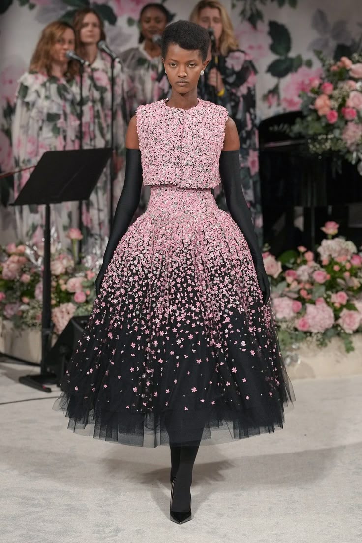 a model stands on a runway wearing a pink and black dress with a floral pattern. behind her are singers on a stage decorated with flowers. she is wearing long black gloves.