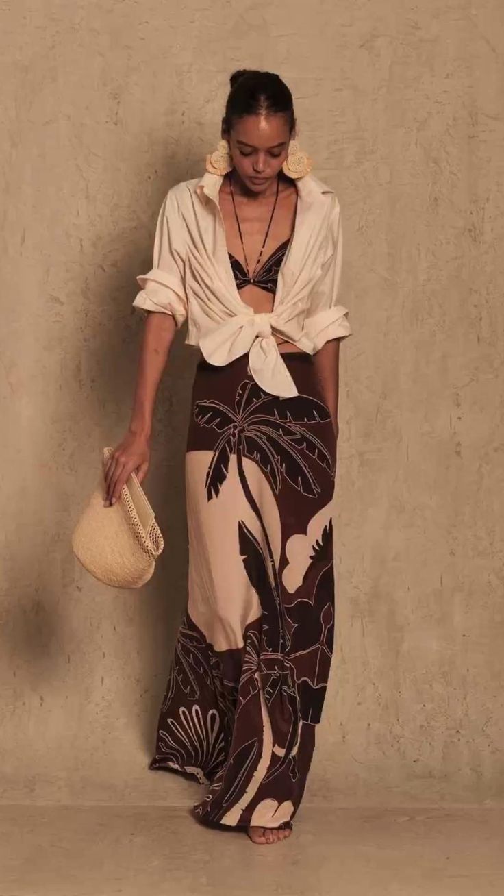 a fullbody shot of a fashion model standing against a textured light beige background. she's wearing a white oversized shirt tied at the waist, a dark brown and beige maxi skirt with a tropical palm tree print, a darkcolored bralette, large beige earrings, and a delicate necklace. she holds a straw handbag in her left hand. the image is welllit and emphasizes the model's attire and accessories. the style is casualchic, summery and beachy, with a slightly bohemian feel.