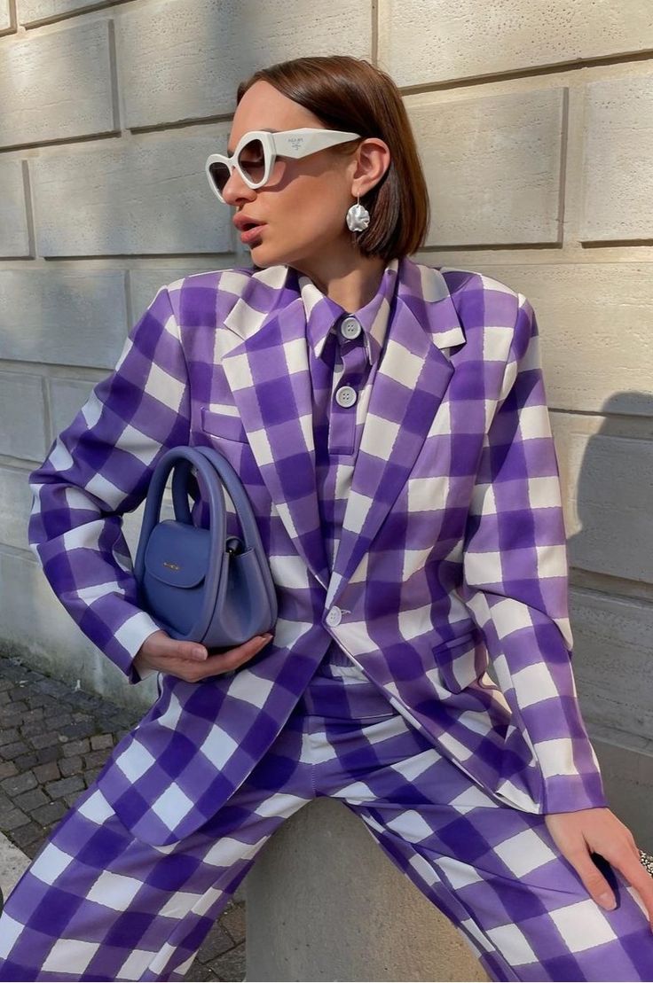 a fashionable woman is posing outdoors, wearing a chic purple and white gingham pantsuit.  she's accessorized with white cateye sunglasses, pearl earrings, and a small light blue handbag. the setting appears to be a sunny day on a city street.  the image focuses on the woman's stylish outfit and accessories.