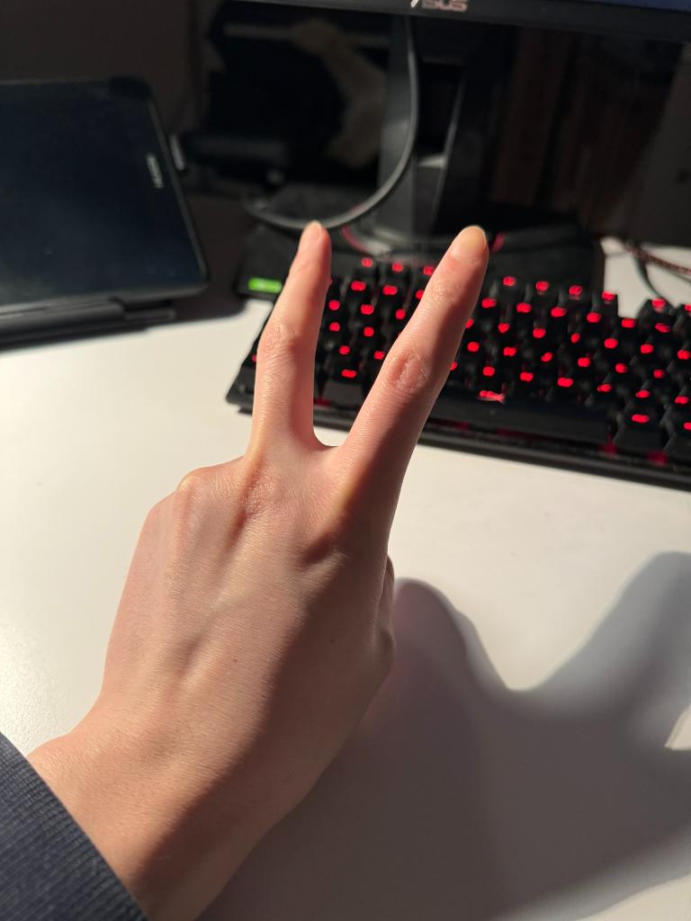 a right hand is making a peace sign in front of a computer desk. there is a black laptop and a keyboard with red backlight on the desk. the photo is taken from a firstperson perspective with artificial lighting.