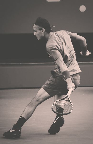 a male tennis player is concentrating as he swings his racket to hit a ball in a tennis match. he is wearing a white shirt with a headband and black shorts. his face is partially obscured by the racket.  he is on the court in an indoor tennis facility.  the photo is in black and white.  he is a powerful athlete, who is clearly focused on hitting the ball with precision.