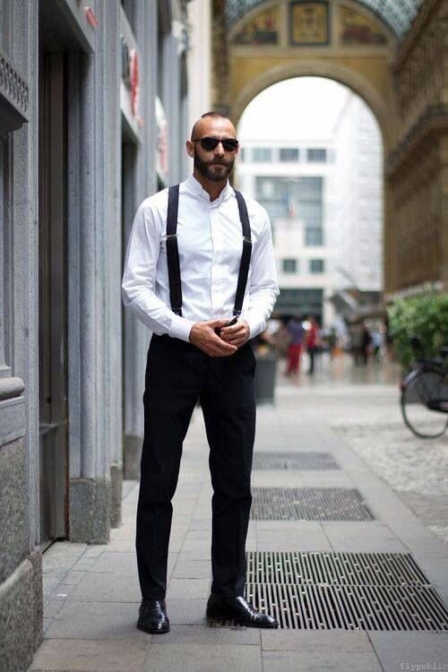 a stylish bald man with sunglasses is standing on a city sidewalk. he is wearing a white buttondown shirt, black suspenders, black dress pants, and black dress shoes. the image is taken from an eyelevel perspective and features natural daylight.  the background shows a city street with buildings and people walking.