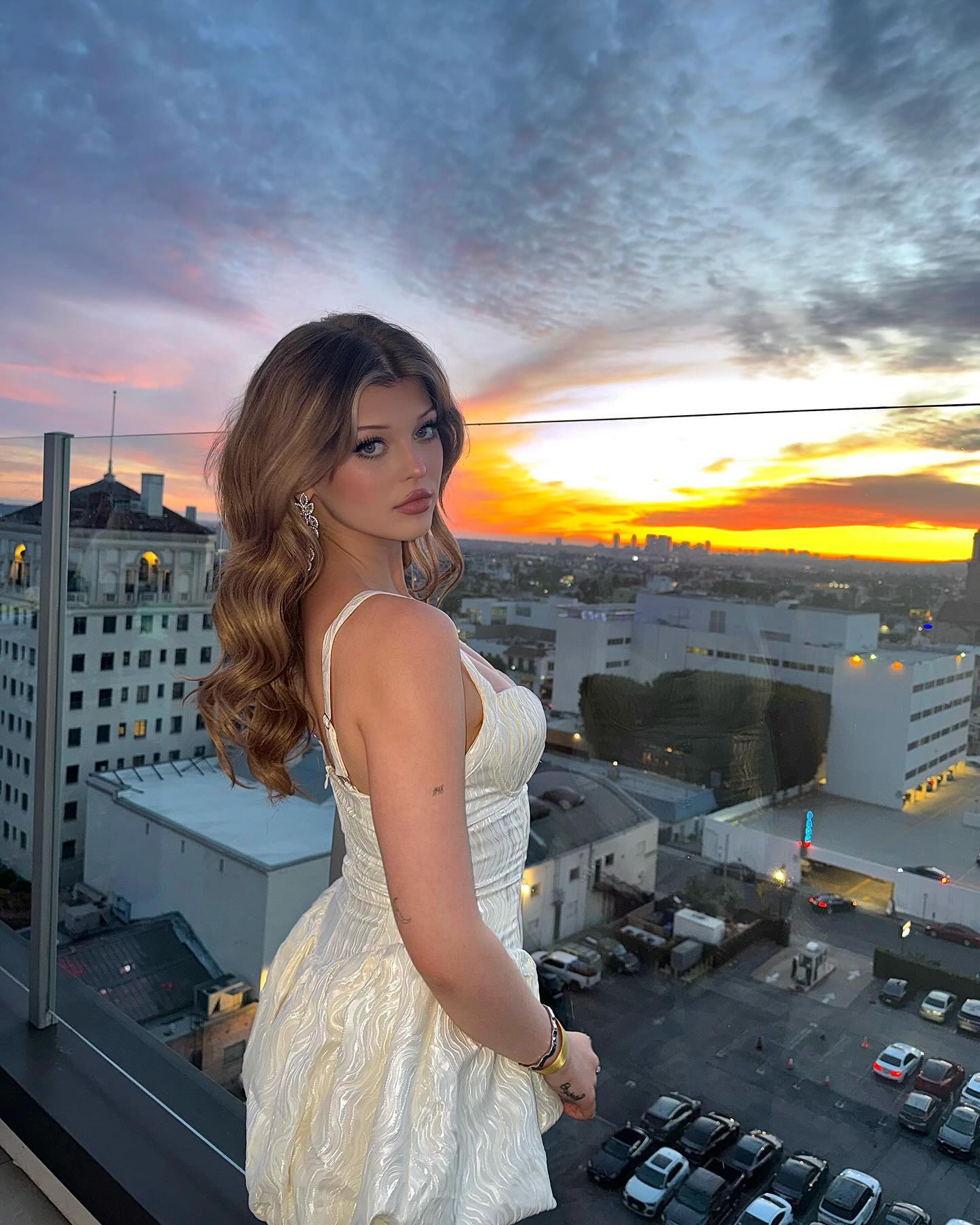 a woman is standing on a rooftop with a cityscape and sunset behind her. she is wearing a white dress and jewelry. the skyline of los angeles is visible in the background.