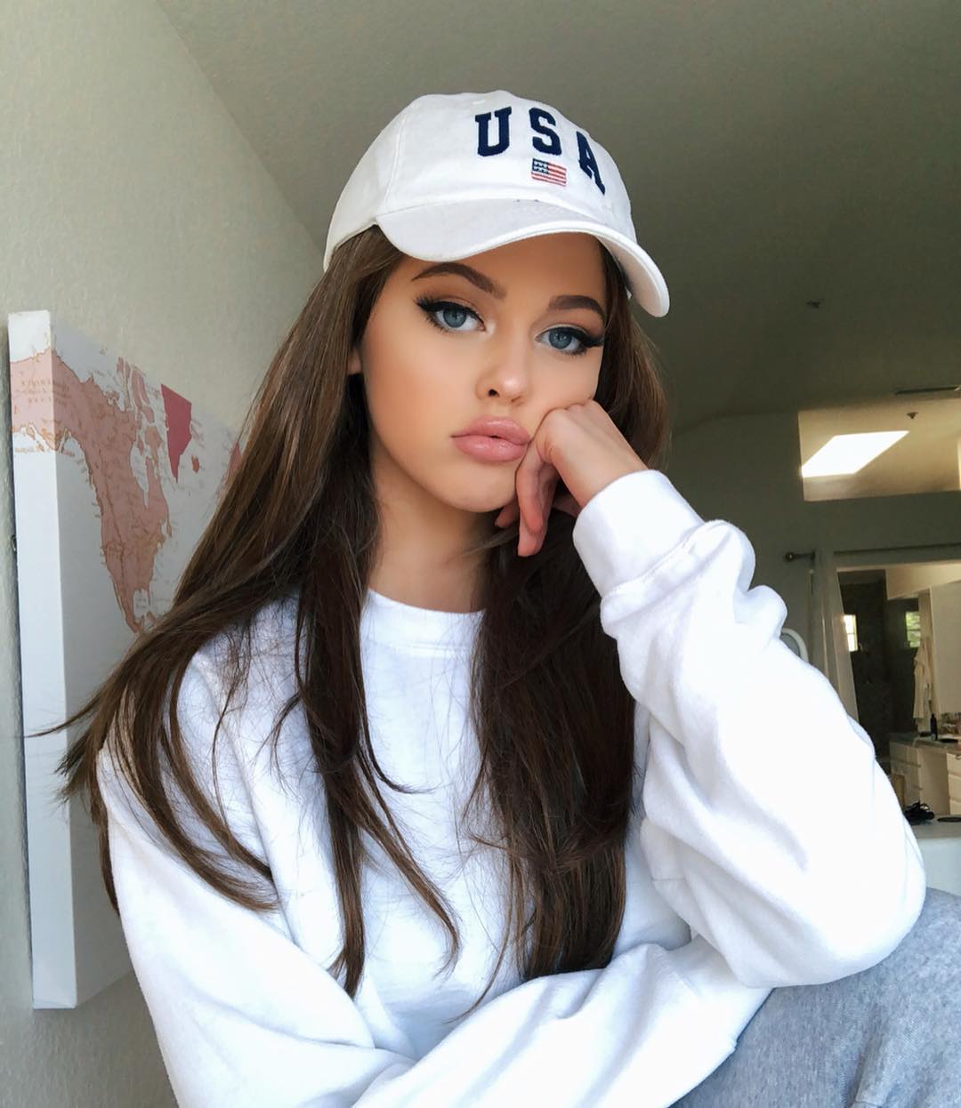 a young woman is sitting and posing for a picture. she's wearing a white sweatshirt, and a white baseball cap that says "usa" on it. her brown hair flows down to her chest. she has blue eyes and is wearing makeup. in the background is a map on the wall.