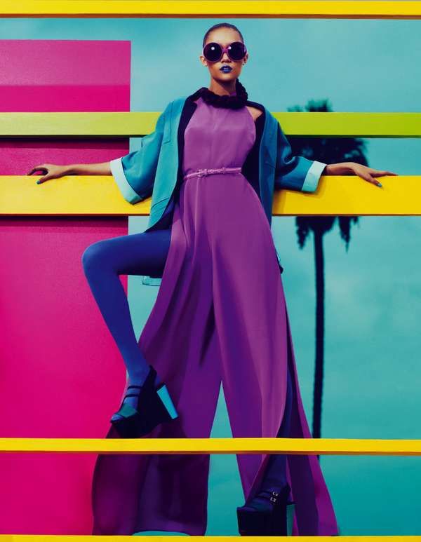 a model in a vibrant fashion shoot poses confidently. she is wearing a purple dress with a turquoise jacket, oversized sunglasses, and striking blue lipstick. she is also wearing platform shoes and blue tights. the backdrop features bold pink, yellow, and turquoise colors, arranged in a gridlike pattern. a palm tree is visible in the background, suggesting a tropical or californian setting. the image is a studio shot.