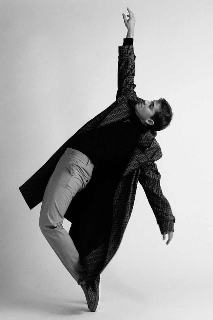 a black and white fullbody shot of a male model in a stylish pose. he is wearing a patterned coat, lightcolored pants, and dark shoes. his arm is raised elegantly, and he appears to be in midmovement, suggesting grace and dynamism. the background is a plain, lightcolored studio setting, focusing attention on the model and his attire. the overall aesthetic is modern, artistic, and highfashion.
