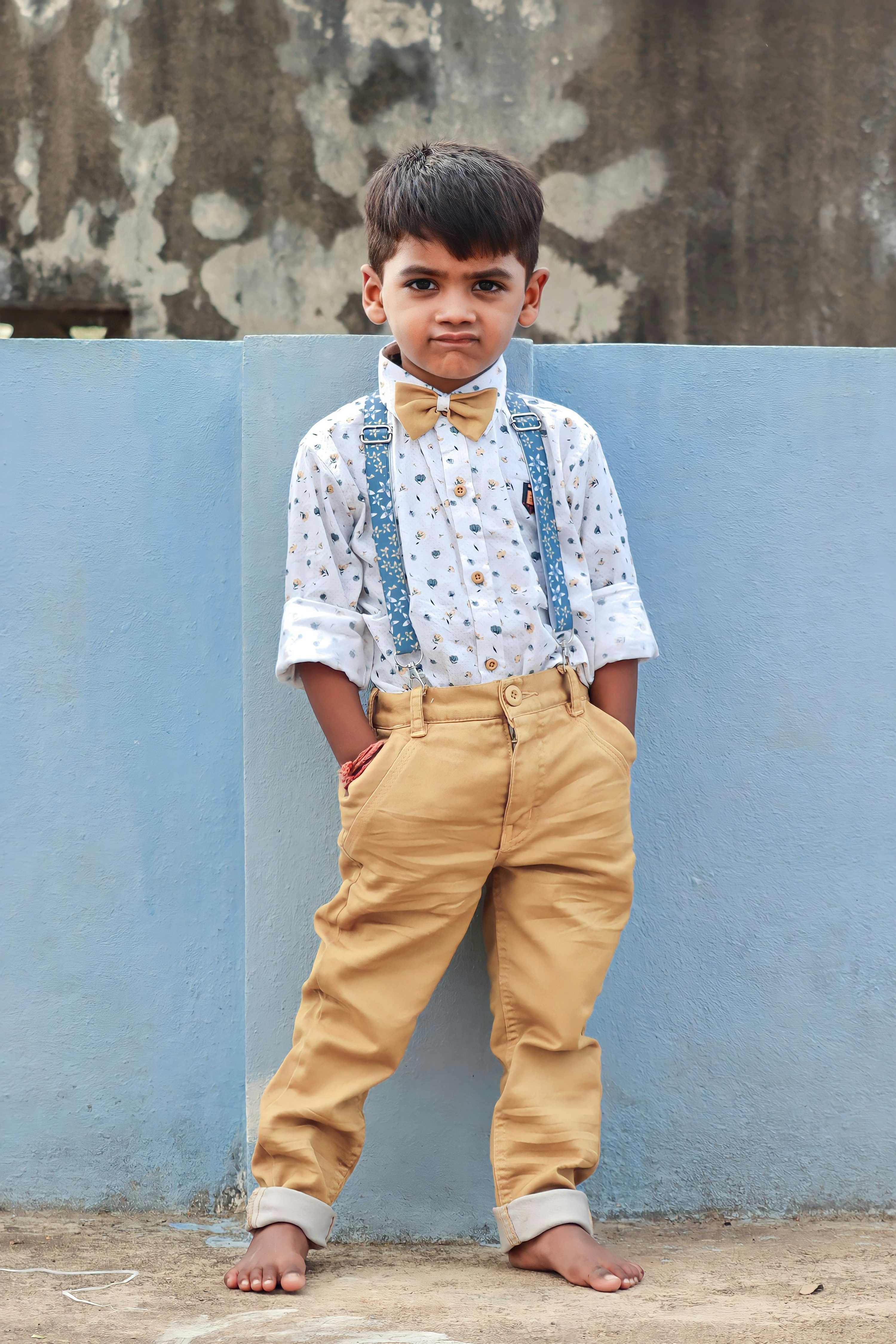 a young boy dressed in formal wear stands barefoot outdoors against a wall. he is wearing a white collared shirt with a pattern, suspenders, a bow tie, and tan pants. he has his hands in his pockets and looks directly at the camera.