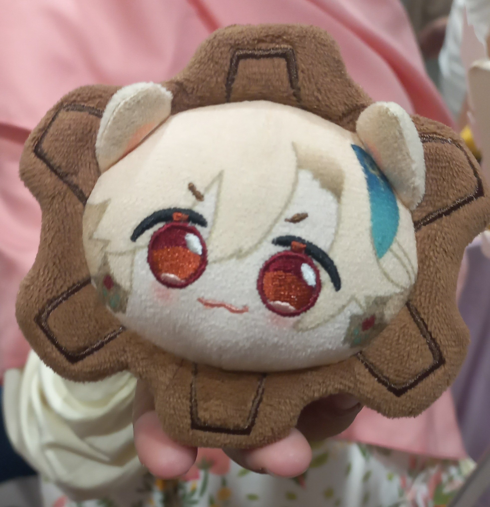 this is a closeup image of a plush toy of a cartoon character with big eyes. the plush is a pale yellow color with brown accents on the face and body. the character has large, orange eyes and brown hair. the plush is being held in a person's hands. the background is a pale pink and yellow color. the photo is taken from a slightly low angle.