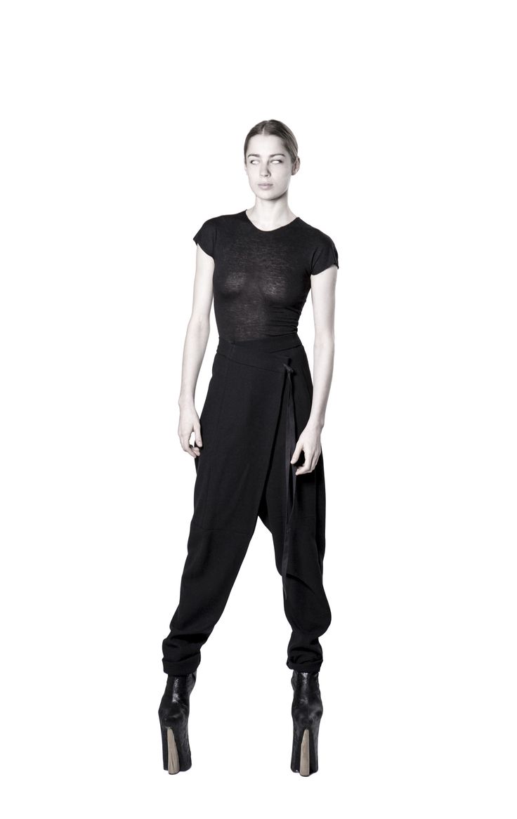 a full shot of a woman in a black and white studio shot. she is wearing a black top and black wrap pants with a belt. she is wearing black boots. she is looking to the side.