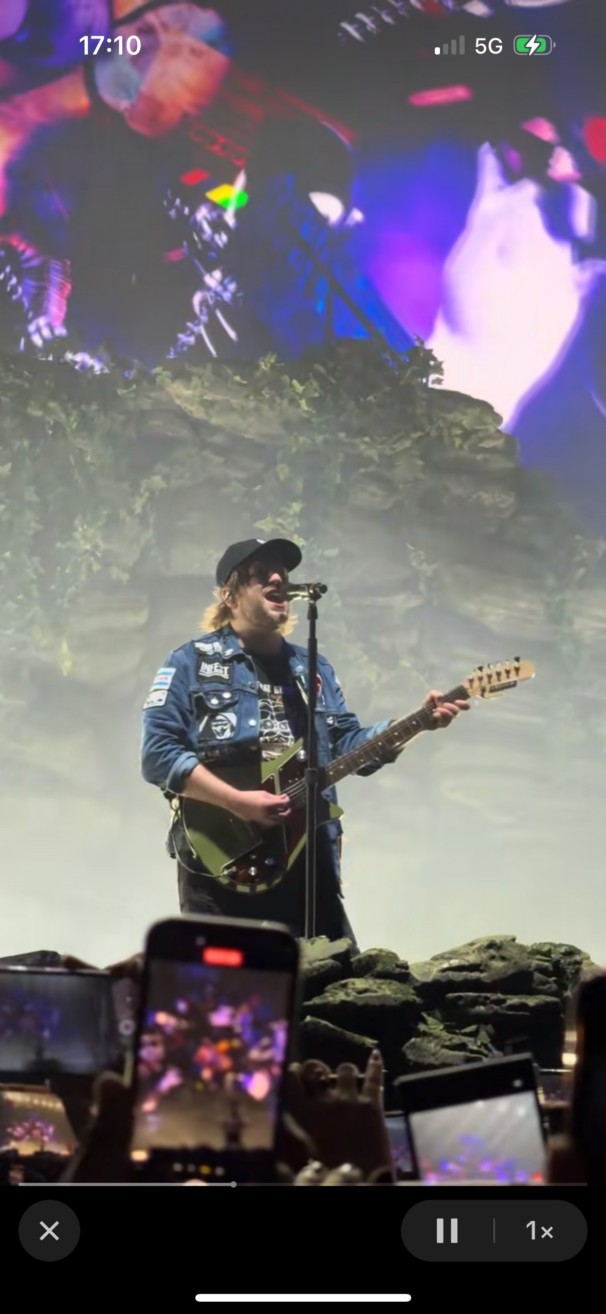 a man in a blue jean jacket stands on stage with a guitar and sings into a microphone while a crowd holds up their phones in the background.