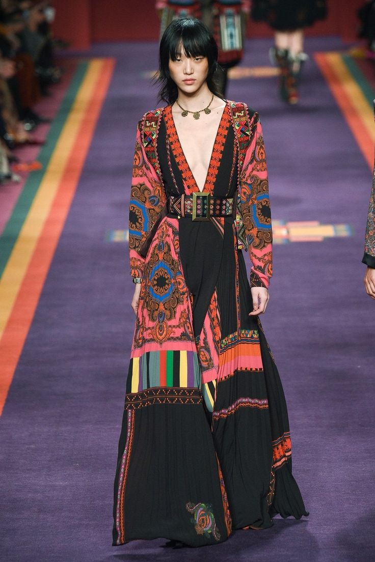 a full shot of a fashion model walking down a runway. she is wearing a long, flowing maxi dress with a vibrant paisley and multicolored print. the dress features a plunging neckline and a fitted bodice, cinched with a wide belt. the skirt is pleated, and the overall style is bohemian chic. the model also wears a gold necklace. the runway is a deep purple, and there are blurred figures in the background. the overall mood is sophisticated and elegant.